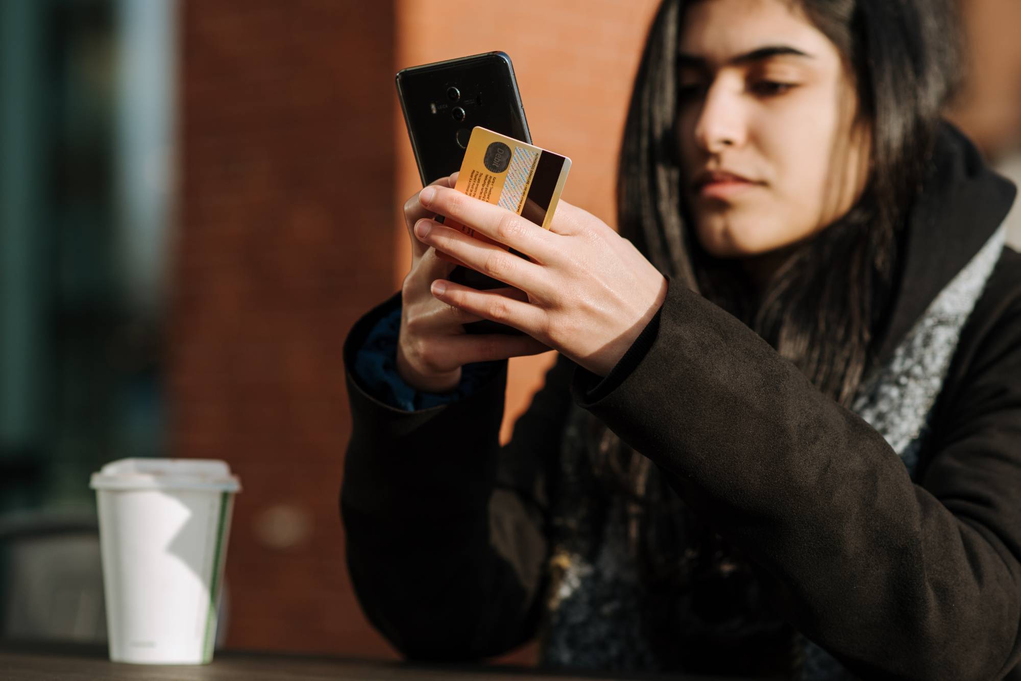 Student performing cashless transaction on mobile phone outside with coffee cup on table